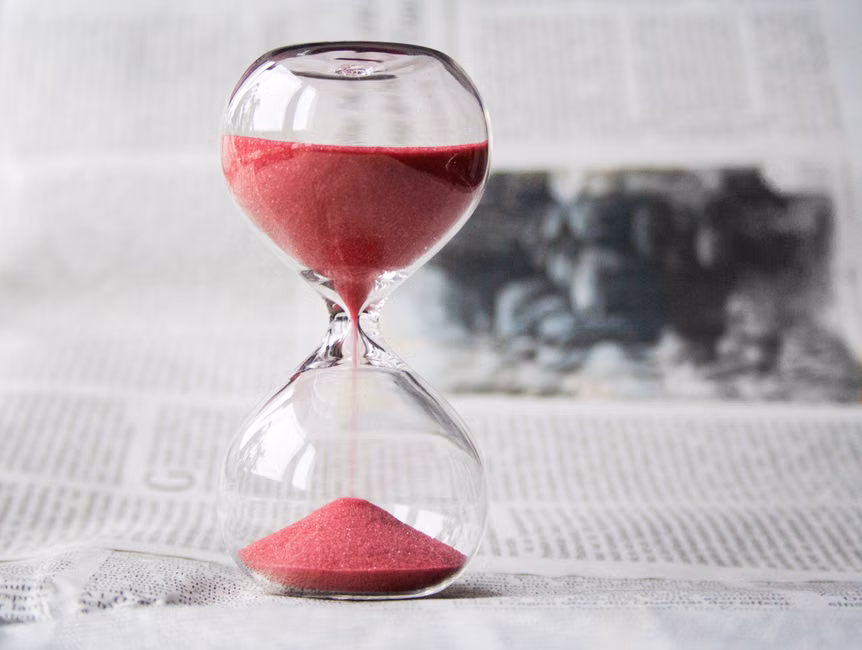 Close-up of a transparent hourglass with pink sand flowing, placed on a newspaper background.